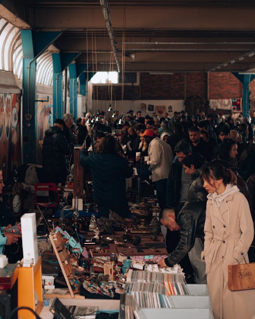 Inside a large indoor market or fairground with high, curved ceilings supported by blue steel beams and exposed brick walls, a diverse crowd of shoppers and vendors browse and inspect various items on numerous tables and display stands. The tables are densely packed with a range of goods including books, antiques, small household items, and decorative objects, some stacked neatly while others are scattered across the surfaces. The lighting is dim with natural light filtering in from windows near the ceiling, complemented by overhead industrial-style lights. In the foreground, a woman in a beige trench coat and a man in a dark jacket are closely examining items on a table, while other individuals on either side browse, hold objects, or engage in conversations. The scene suggests a marketplace or second-hand sale event, which may involve independent collection or disposal of a variety of items, consistent with private waste handling or rubbish clearance services like those offered by Rubbish Clearance Shepherds Bush.
