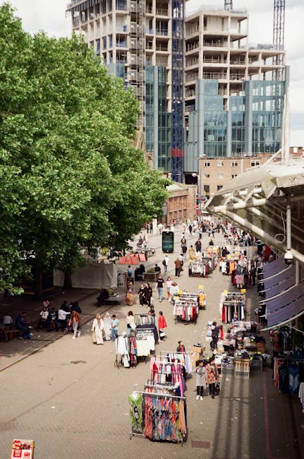 A city street scene featuring a mix of historic and modern multi-storey buildings with brick and stone facades, large sash windows, and decorative architectural details. The foreground shows a white rubbish collection vehicle parked near an entrance with stone pillars and greenery, indicating activity related to waste clearance or disposal. The street is lined with bollards and bicycles parked along the pavement, while other vehicles, including a bus, are visible further down the road. The lighting suggests daytime with overcast skies, creating a soft, diffused atmosphere. The context of rubbish removal services is subtly implied by the presence of the collection vehicle and waste handling activities in an urban setting, aligning with alternative waste management approaches outside of regular municipal collection. These detailed features reflect a typical environment where independent rubbish clearance by companies like [COMPANY_NAME] might operate to serve local businesses or residents in need of private waste disposal solutions.