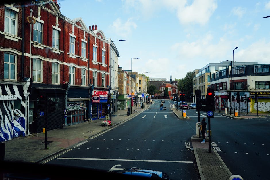 A wide view of a busy urban street scene shows a row of multi-story brick and modern buildings lining the left side of the road, with various shops and storefronts visible at street level, including a business with a sign reading 'CARINA' and graffiti-style artwork on a shutter. The right side features modern buildings with glass balcony accents, and there are several traffic lights showing red, alongside pedestrian crossings and street poles with bollards. The street has multiple lanes with directional arrows painted on the asphalt, and a few vehicles are visible in the distance, including a car and a van. Pedestrians are waiting at the crosswalks, some standing on the sidewalk, which is equipped with tactile paving and bollards for safety. The scene is illuminated by daylight under a partly cloudy sky, and the environment appears clean and organized, consistent with a city area where private rubbish or waste removal services might operate to maintain cleanliness amidst urban activity, as highlighted by services like rubbishclearanceshepherdsbush.co.uk.