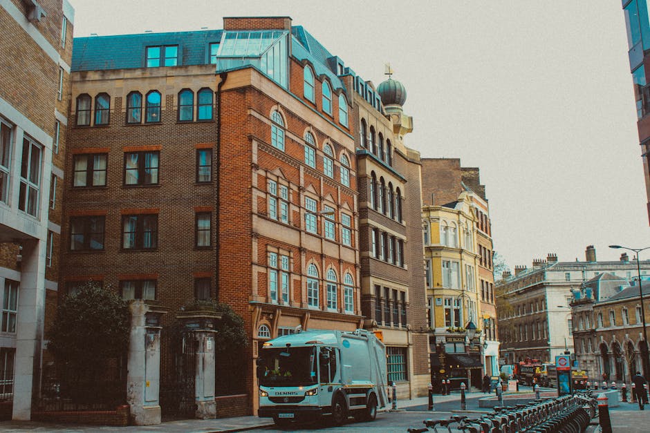 A wide view of a busy urban street scene shows a row of multi-story brick and modern buildings lining the left side of the road, with various shops and storefronts visible at street level, including a business with a sign reading 'CARINA' and graffiti-style artwork on a shutter. The right side features modern buildings with glass balcony accents, and there are several traffic lights showing red, alongside pedestrian crossings and street poles with bollards. The street has multiple lanes with directional arrows painted on the asphalt, and a few vehicles are visible in the distance, including a car and a van. Pedestrians are waiting at the crosswalks, some standing on the sidewalk, which is equipped with tactile paving and bollards for safety. The scene is illuminated by daylight under a partly cloudy sky, and the environment appears clean and organized, consistent with a city area where private rubbish or waste removal services might operate to maintain cleanliness amidst urban activity, as highlighted by services like rubbishclearanceshepherdsbush.co.uk.