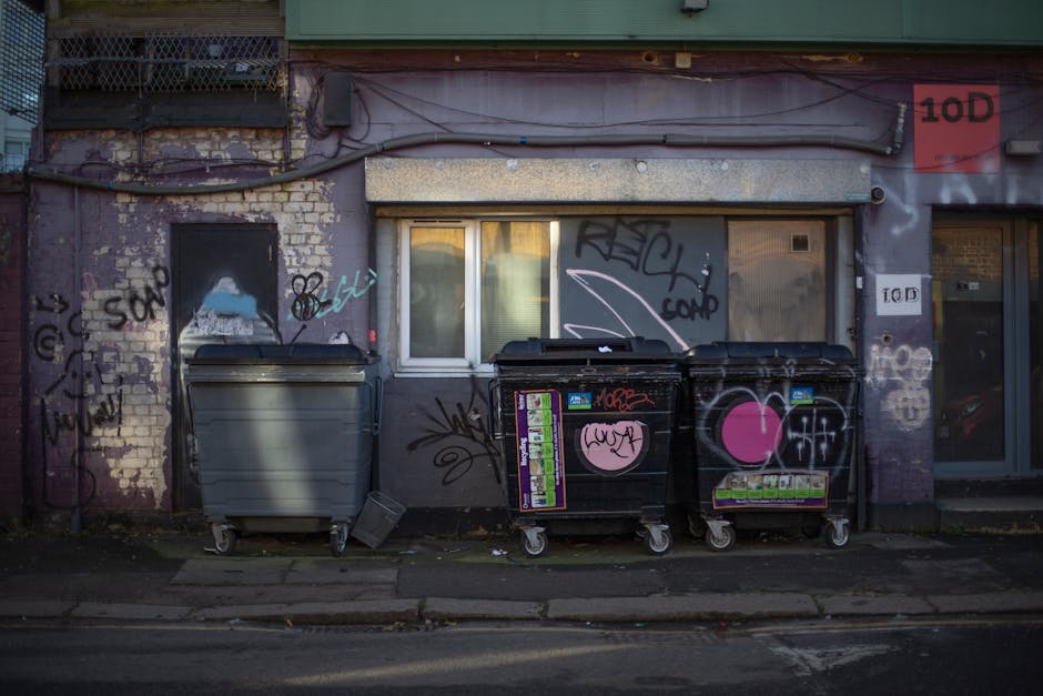 Three large commercial rubbish bins are positioned side by side on a concrete sidewalk beneath a weathered building facade. The bins consist of one grey bin on the left and two black bins on the right, all with sturdy plastic bodies, wheels, and closed lids. The black bins are decorated with various pieces of graffiti, including a large pink heart, scribbled tags, and stickers, contrasting with the plain grey bin. The background features a purple-painted brick wall with visible signs of age, peeling paint, and graffiti tags, above which runs an exposed conduit pipe and electrical wiring. Behind the bins, there's a window with closed blinds, and a metal security shutter partially rolled down, both integrated into the building’s facade, which exhibits grime, chipped paint, and additional graffiti. The scene captures an urban environment where private waste collection or alternative rubbish clearance services, such as those provided by rubbishclearanceshepherdsbush.co.uk, might be involved in managing such waste disposal, reflecting routines of street-level rubbish management in Shepherds Bush.